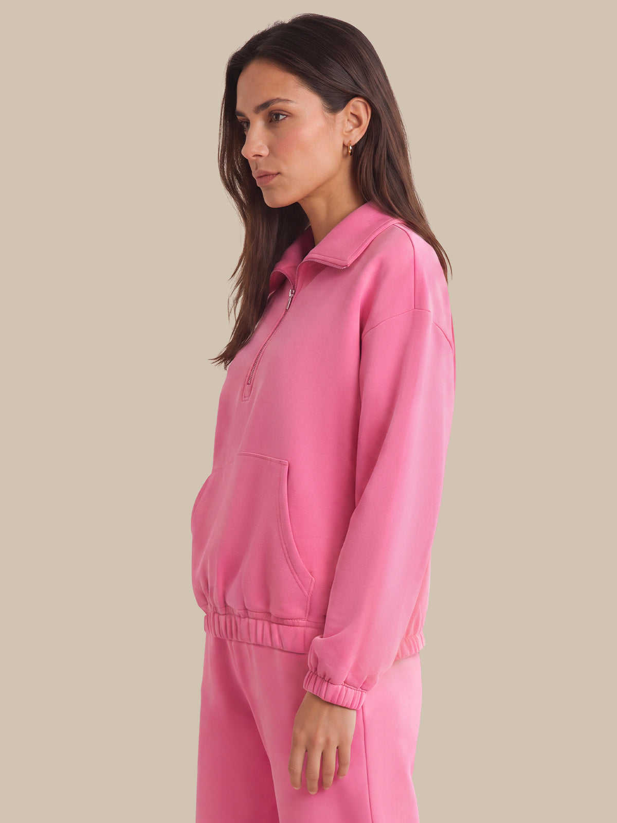A woman wearing a bright pink matching lounge set, featuring a long-sleeve half-zip pullover with a collar and front pouch pocket, paired with matching jogger pants, set against a solid beige background.