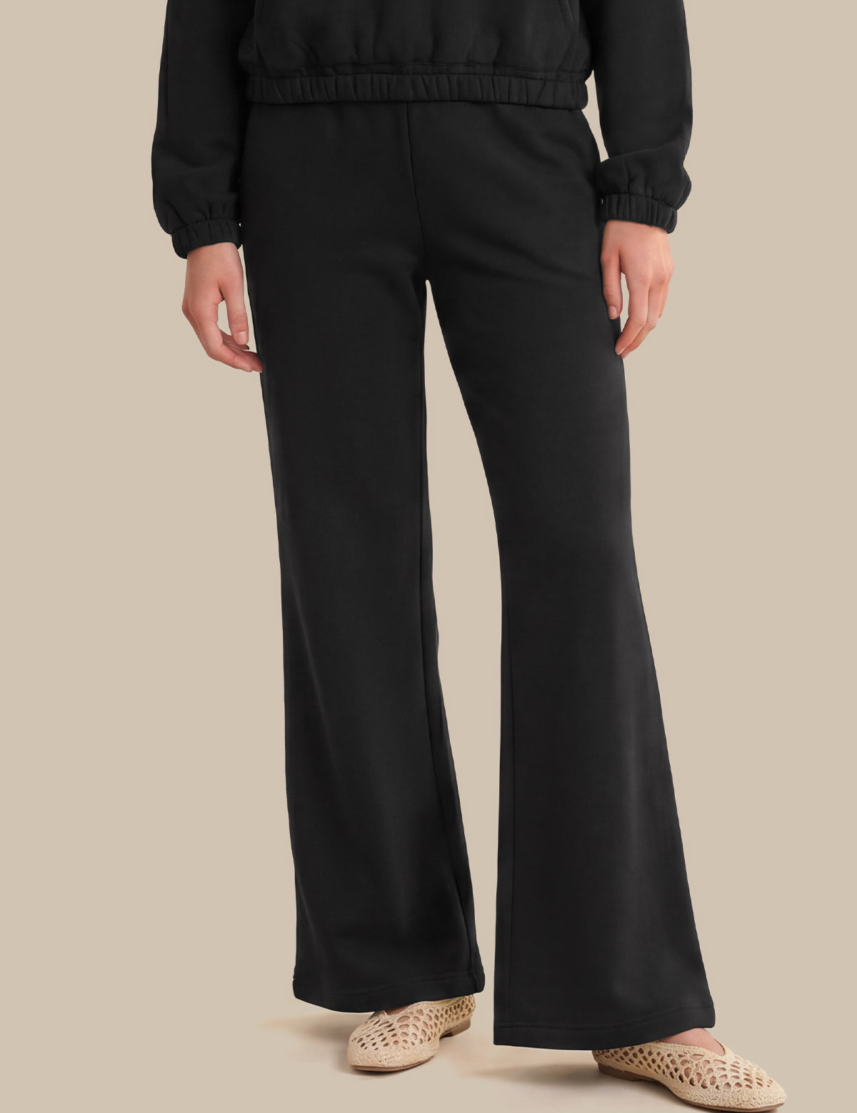A person wearing black wide-leg pants with a matching black cropped jacket, paired with beige woven flat shoes, against a solid beige background.