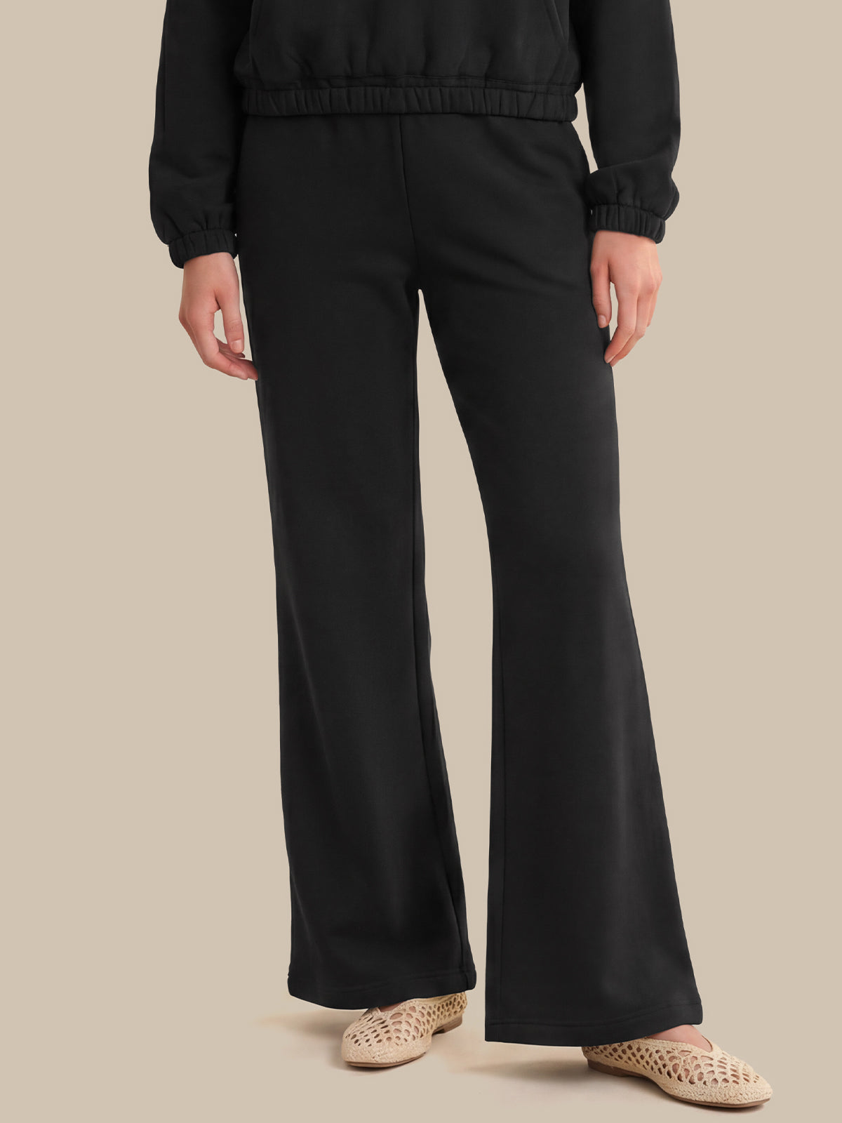 A person wearing black wide-leg pants with a matching black cropped jacket, paired with beige woven flat shoes, against a solid beige background.