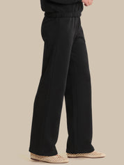 Black wide-leg trousers shown on a model from the waist down, paired with beige woven flat shoes against a solid beige background.