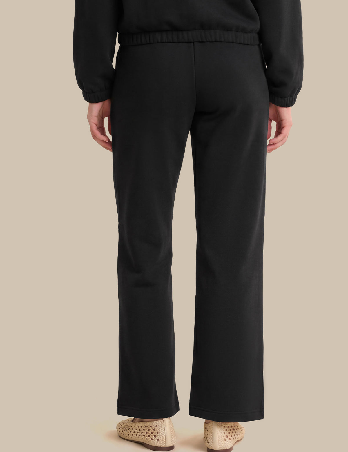 Rear view of a person wearing black wide-leg trousers with a matching black top, paired with beige woven flat shoes, against a solid beige background.