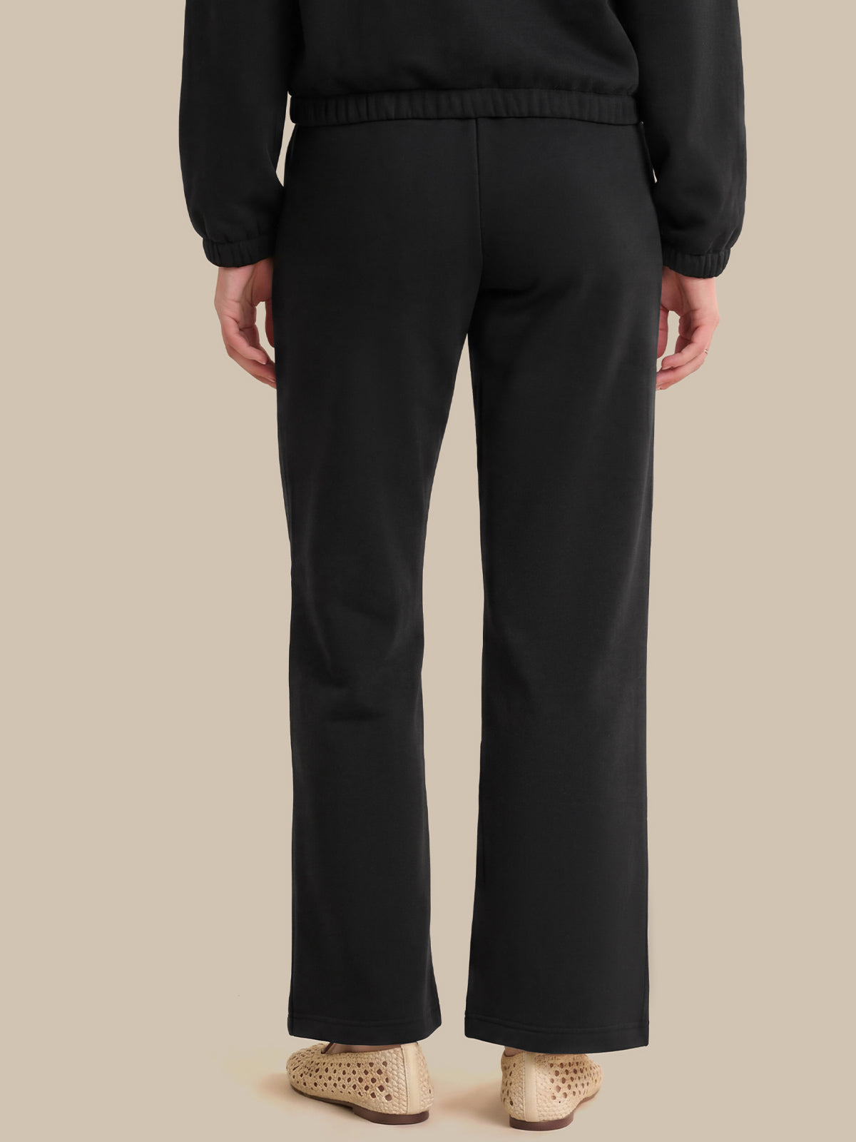 Rear view of a person wearing black wide-leg trousers with a matching black top, paired with beige woven flat shoes, against a solid beige background.