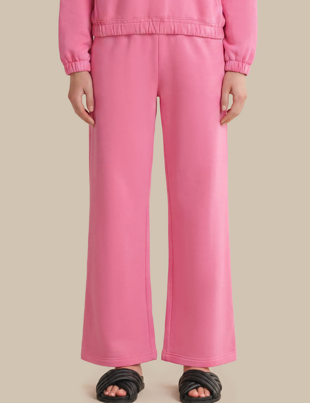 A person wearing bright pink wide-leg sweatpants with a matching top and black slide sandals, shown from the chest down against a plain beige studio background.