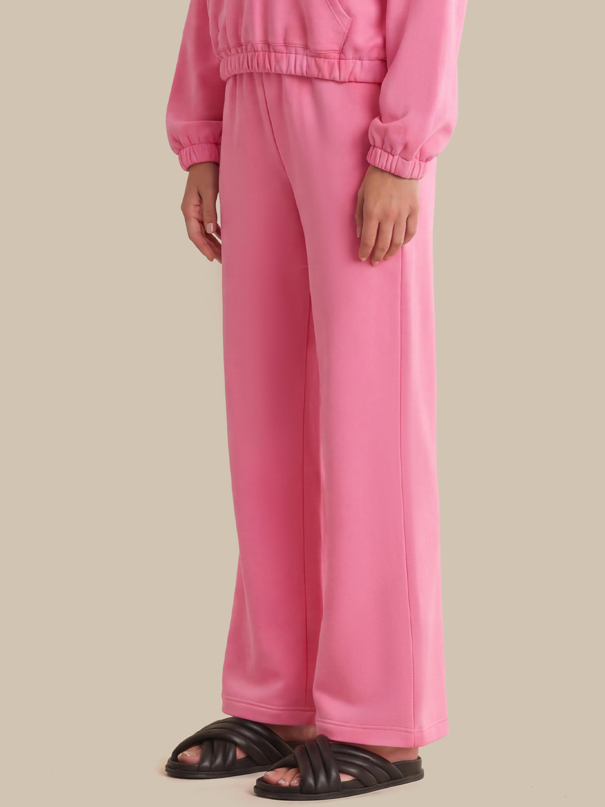 A person wearing a matching pink lounge set with wide-leg sweatpants and a cropped long-sleeve top, paired with black slide sandals, against a plain beige studio background.