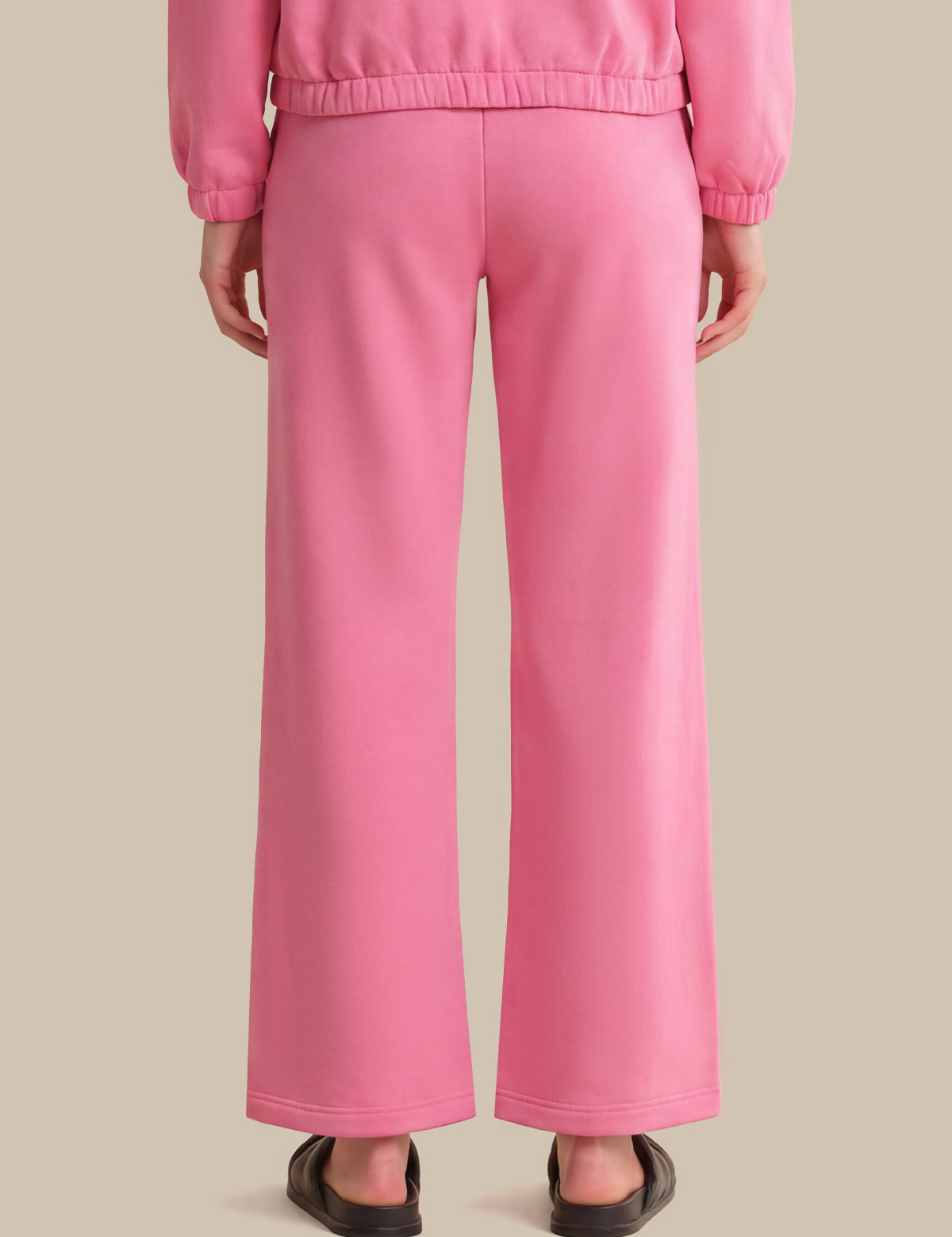 A person shown from the back wearing bright pink wide-leg pants and a matching pink top, standing against a plain warm beige studio background.