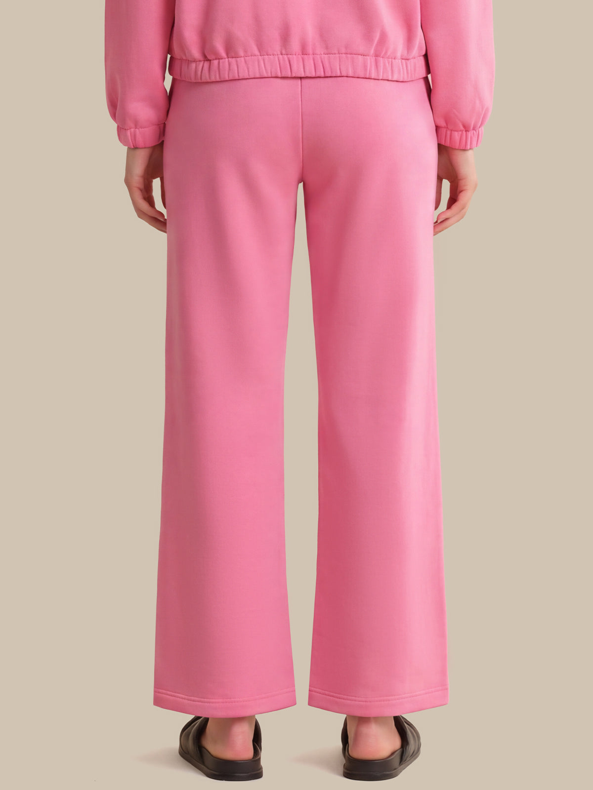 A person shown from the back wearing bright pink wide-leg pants and a matching pink top, standing against a plain warm beige studio background.