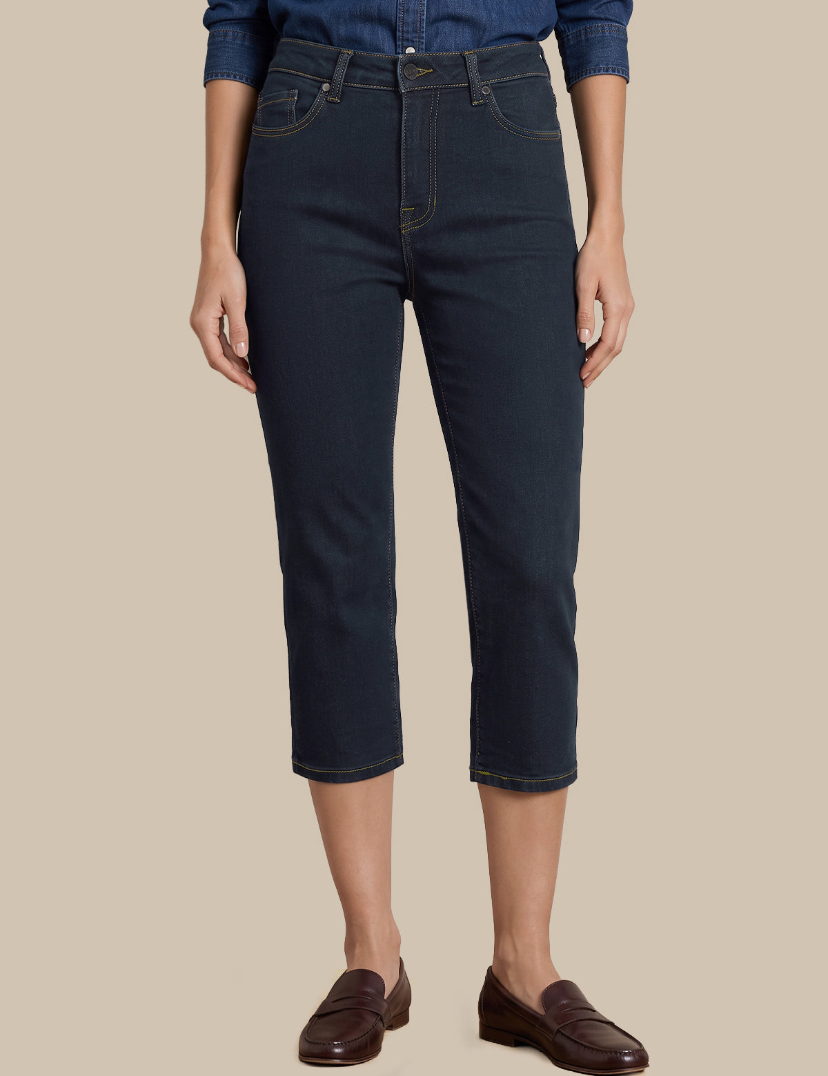 A model wearing dark navy blue cropped jeans, a blue denim shirt, and dark brown leather loafers, shown from the waist down against a solid beige background.