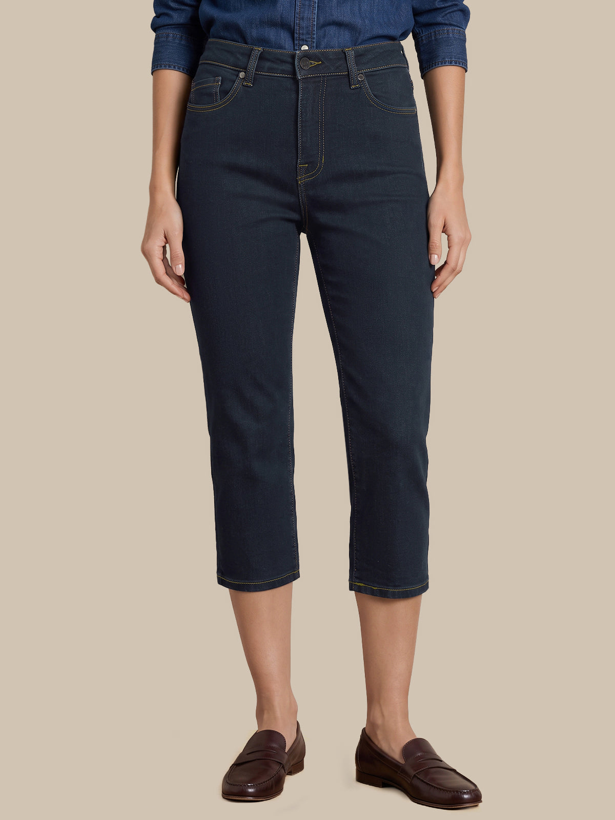 A model wearing dark navy blue cropped jeans, a blue denim shirt, and dark brown leather loafers, shown from the waist down against a solid beige background.