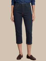 A model wearing dark navy blue cropped jeans, a blue denim shirt, and dark brown leather loafers, shown from the waist down against a solid beige background.