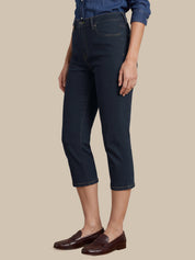 A model wearing dark navy blue cropped denim jeans, a blue denim shirt, and dark brown leather loafers, shown from the waist down against a solid beige background.