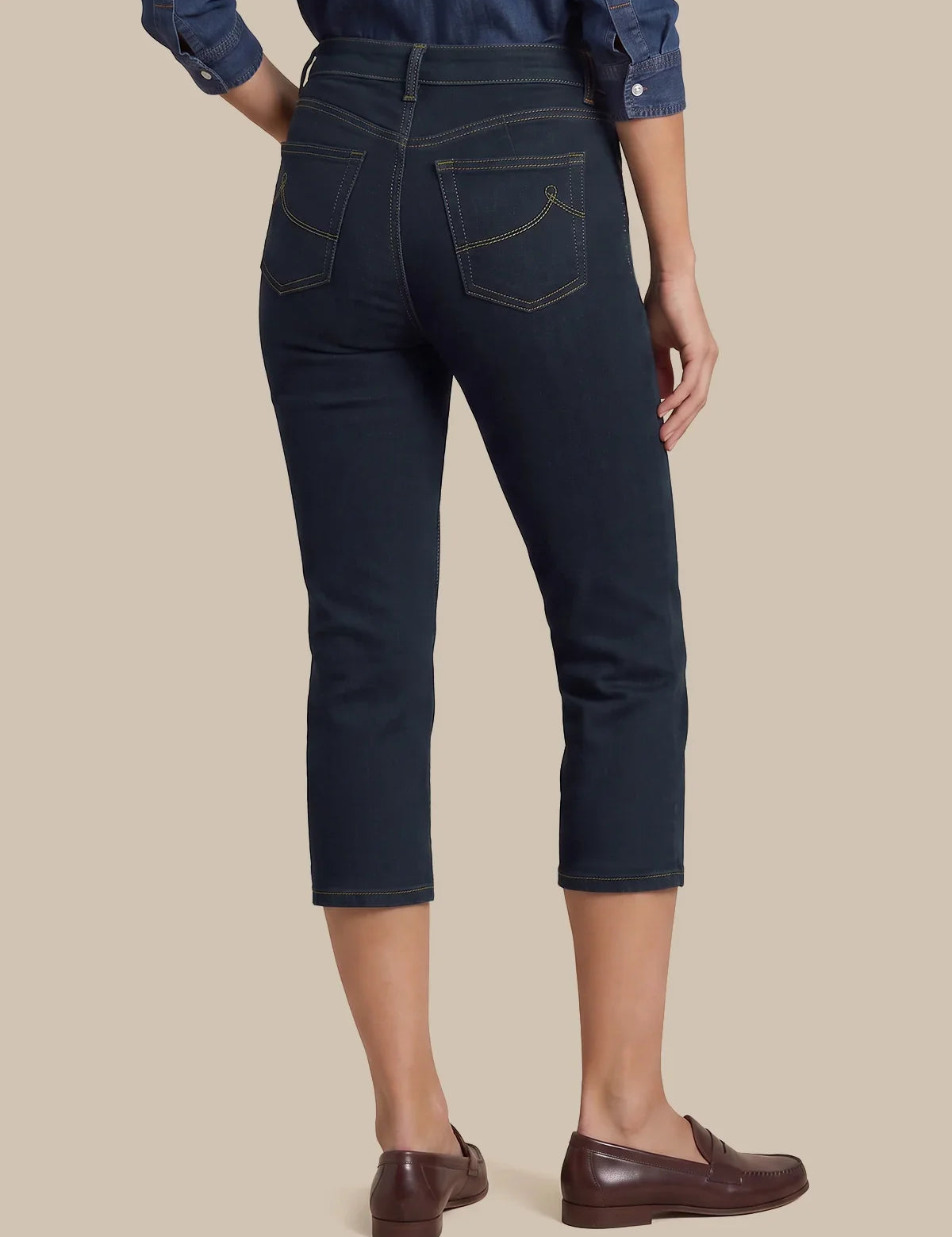 Rear view of a person wearing dark navy cropped jeans with contrast gold stitching, paired with a blue denim shirt and brown leather loafers, against a solid beige background.