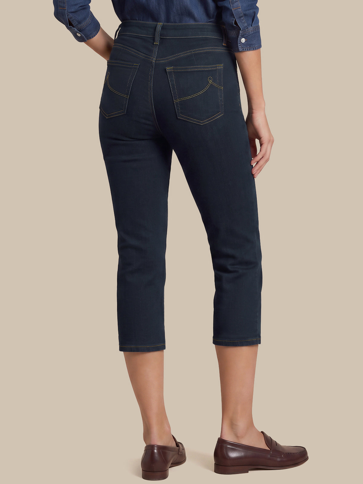 Rear view of a person wearing dark navy cropped jeans with contrast gold stitching, paired with a blue denim shirt and brown leather loafers, against a solid beige background.