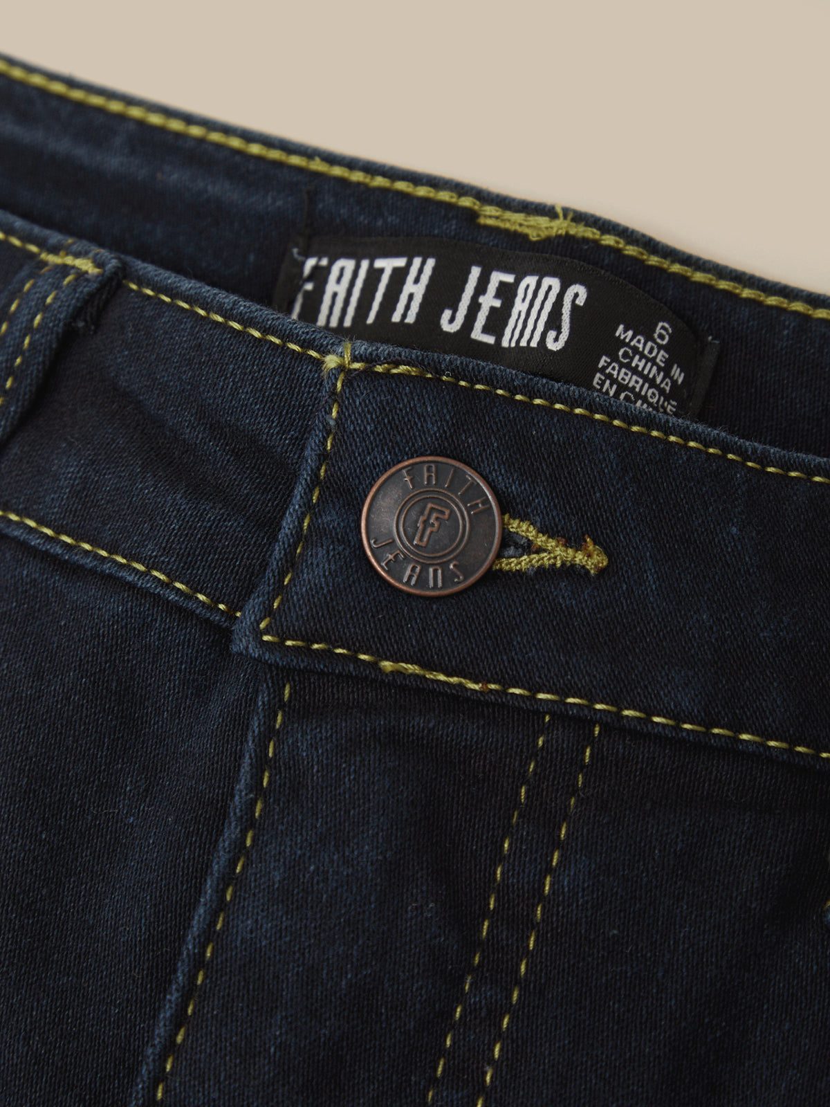 Close-up of dark indigo denim jeans showing the waistband, yellow contrast stitching, a round metal button, and a visible Faith Jeans brand label.