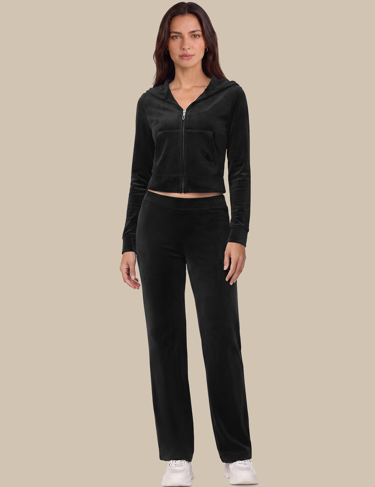 A woman modeling a matching black velour tracksuit with a cropped zip-up hoodie and straight-leg pants, paired with white sneakers, against a solid beige background.
