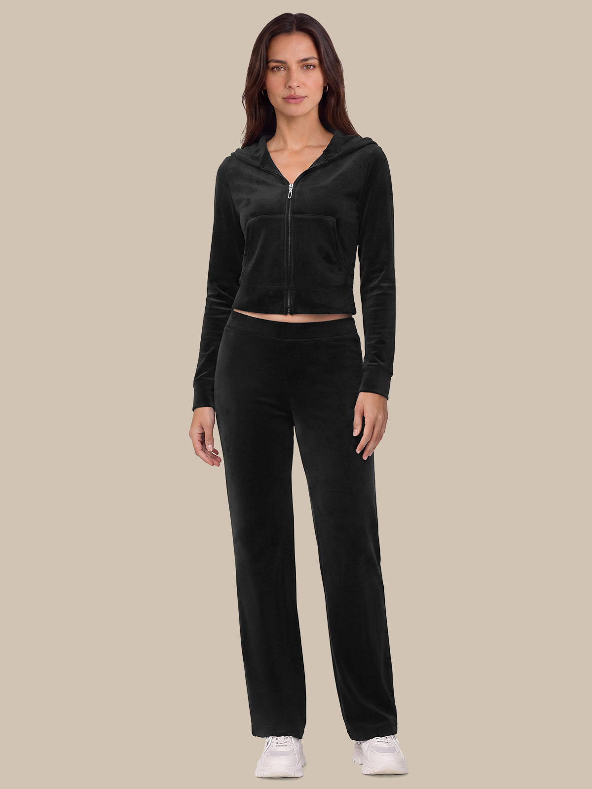 A woman modeling a matching black velour tracksuit with a cropped zip-up hoodie and straight-leg pants, paired with white sneakers, against a solid beige background.