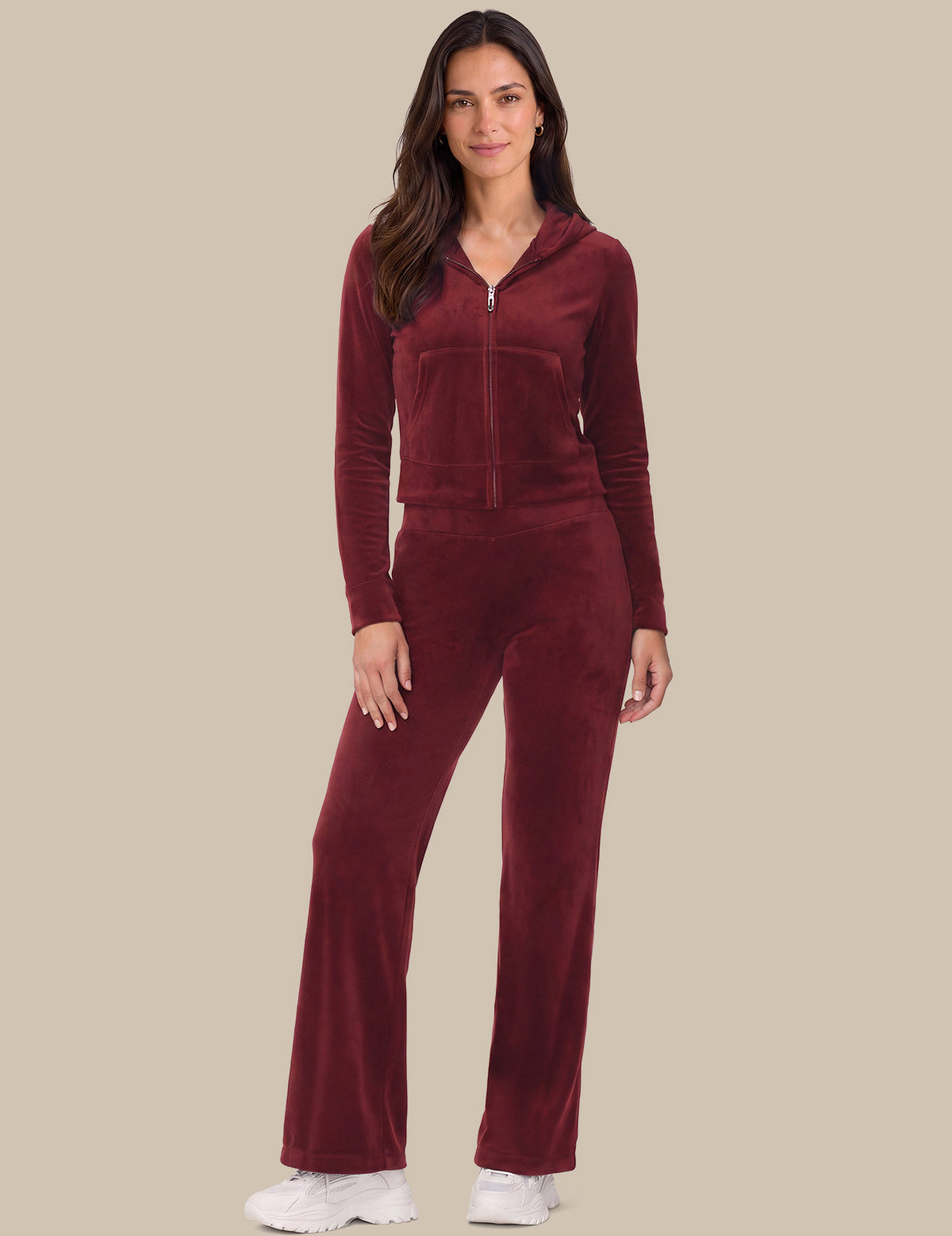 A woman modeling a matching burgundy velour tracksuit with a zip-up hoodie and wide-leg pants, paired with white sneakers, against a solid beige background.