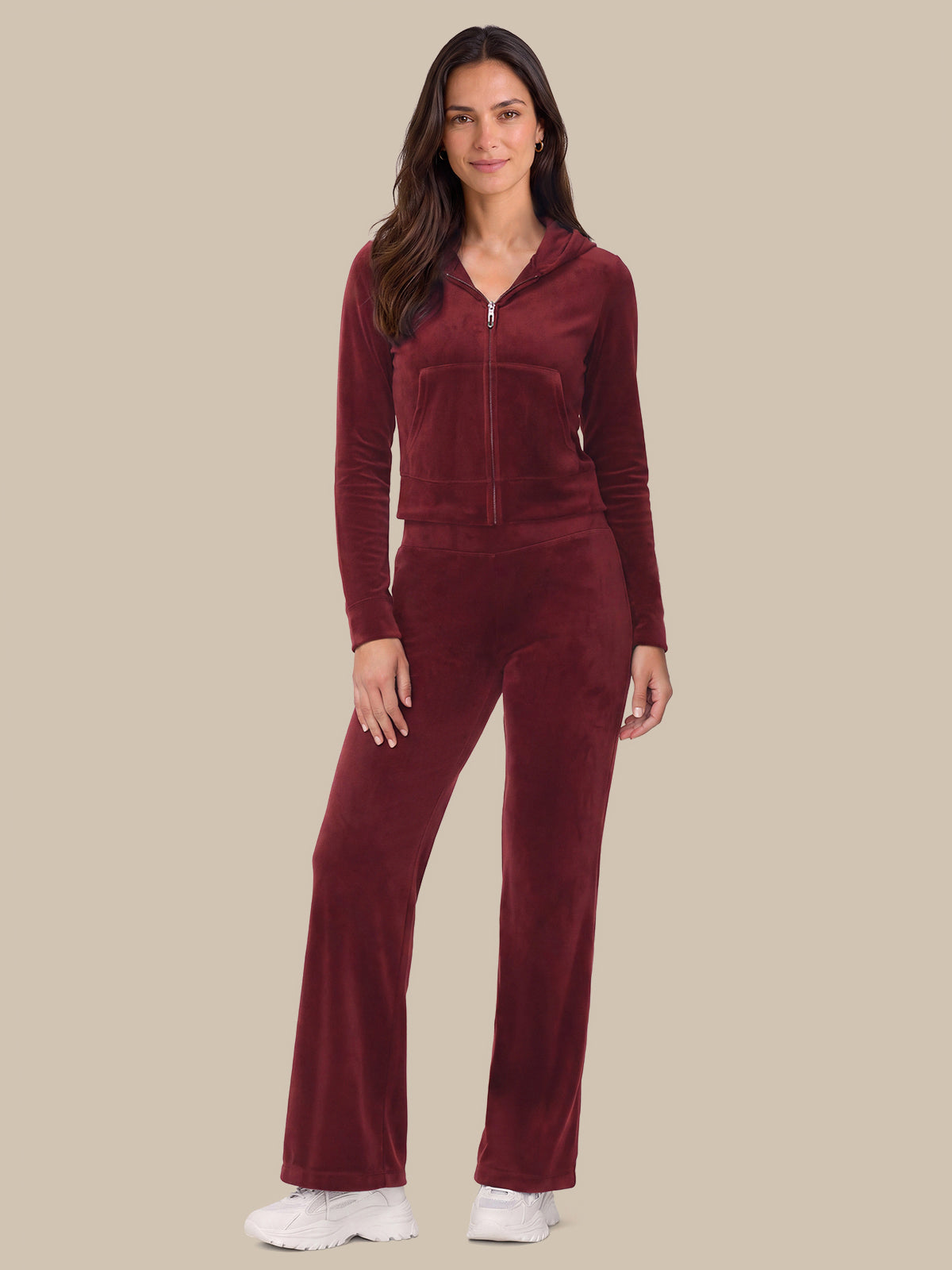 A woman modeling a matching burgundy velour tracksuit with a zip-up hoodie and wide-leg pants, paired with white sneakers, against a solid beige background.