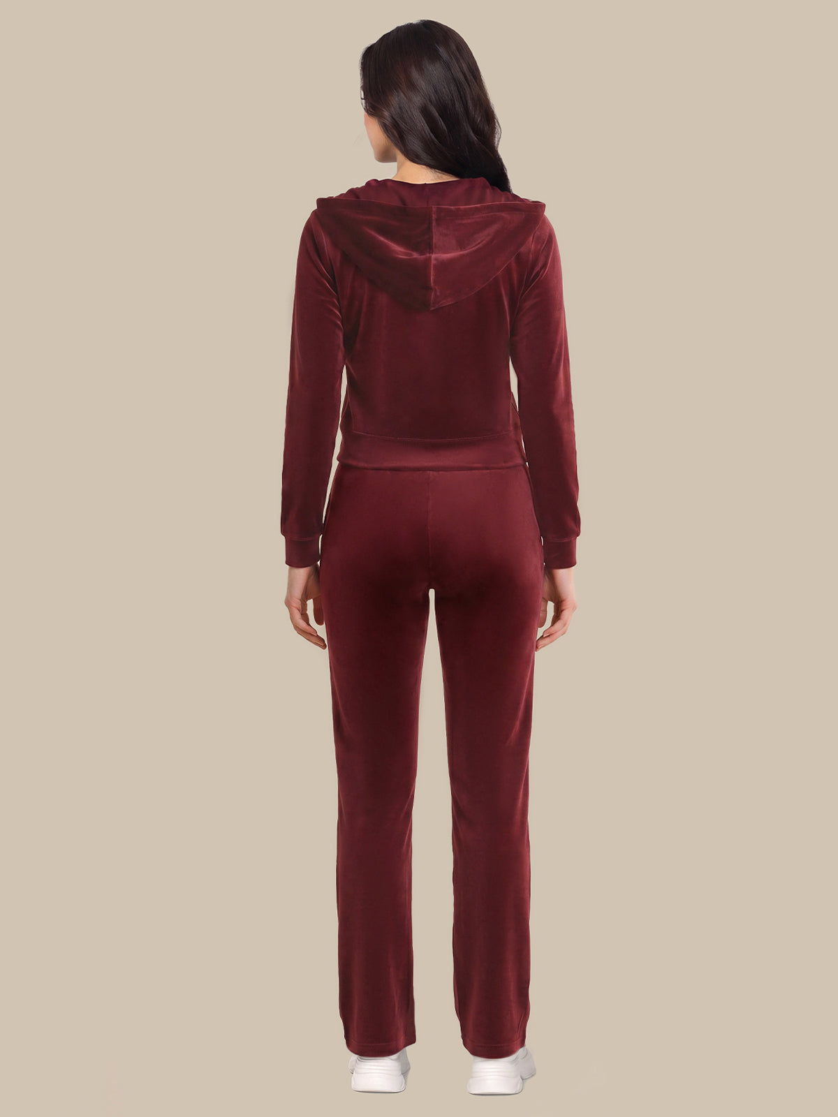 A woman viewed from the back wearing a matching burgundy velvet tracksuit with a hooded long-sleeve top and straight-leg pants, standing against a solid beige background.