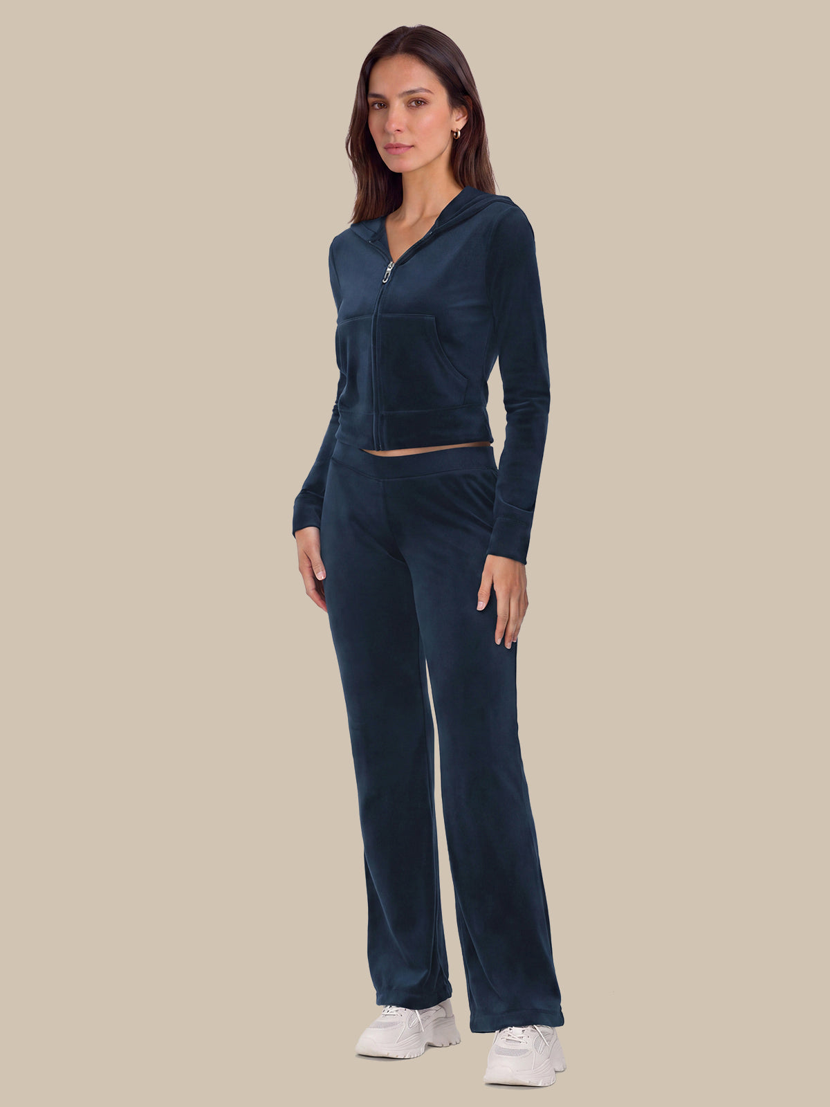 A woman modeling a matching dark navy blue velour tracksuit, consisting of a cropped zip-up hoodie and wide-leg pants, paired with white sneakers, against a solid beige background.