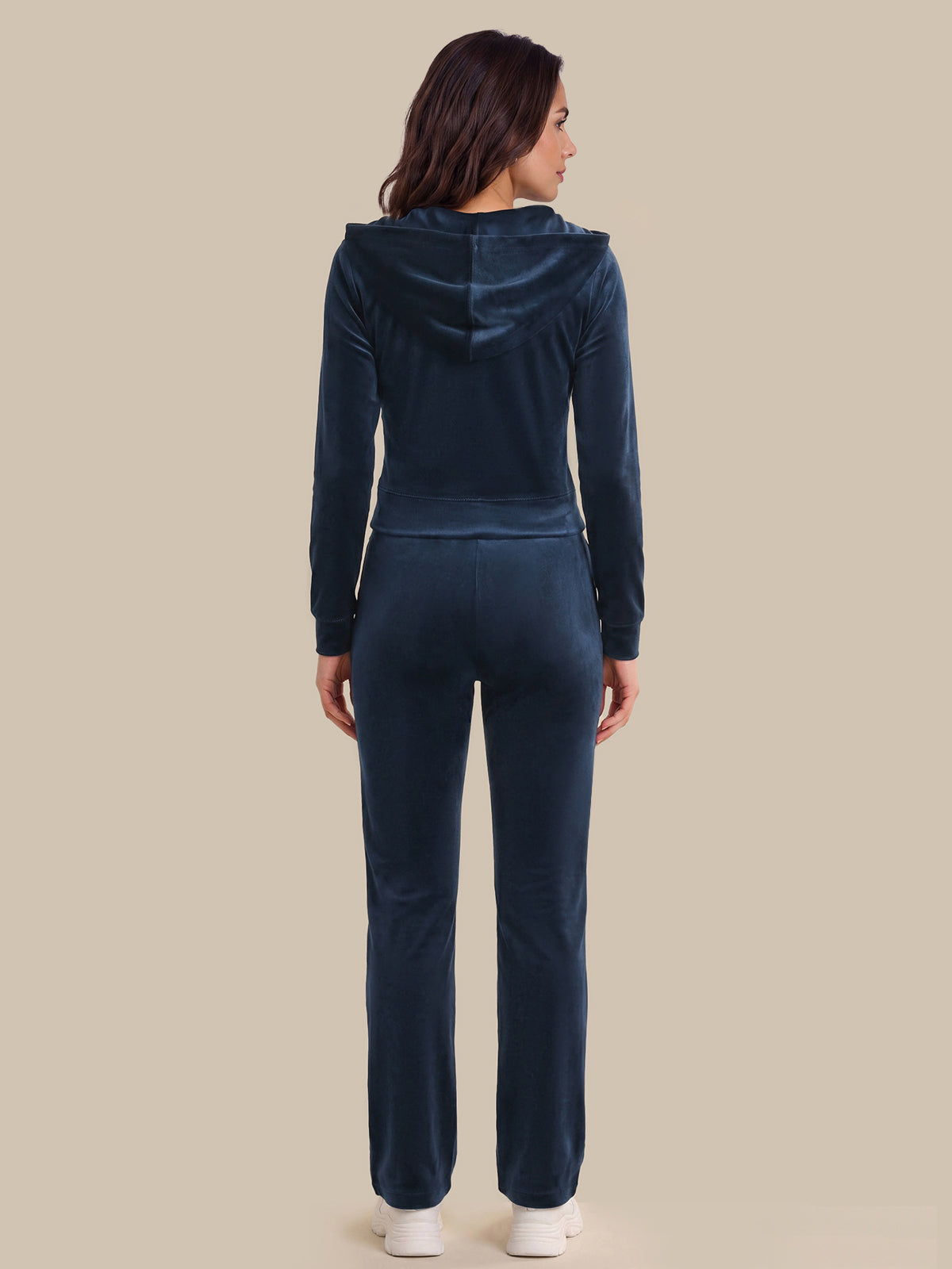A woman viewed from the back wearing a matching dark navy velour tracksuit with a hooded long-sleeve top and straight-leg pants, standing against a solid beige background.