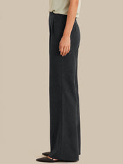 A side-profile view of a model wearing dark charcoal gray wide-leg knit pants, paired with a light beige top and black pointed flats, against a solid beige background.