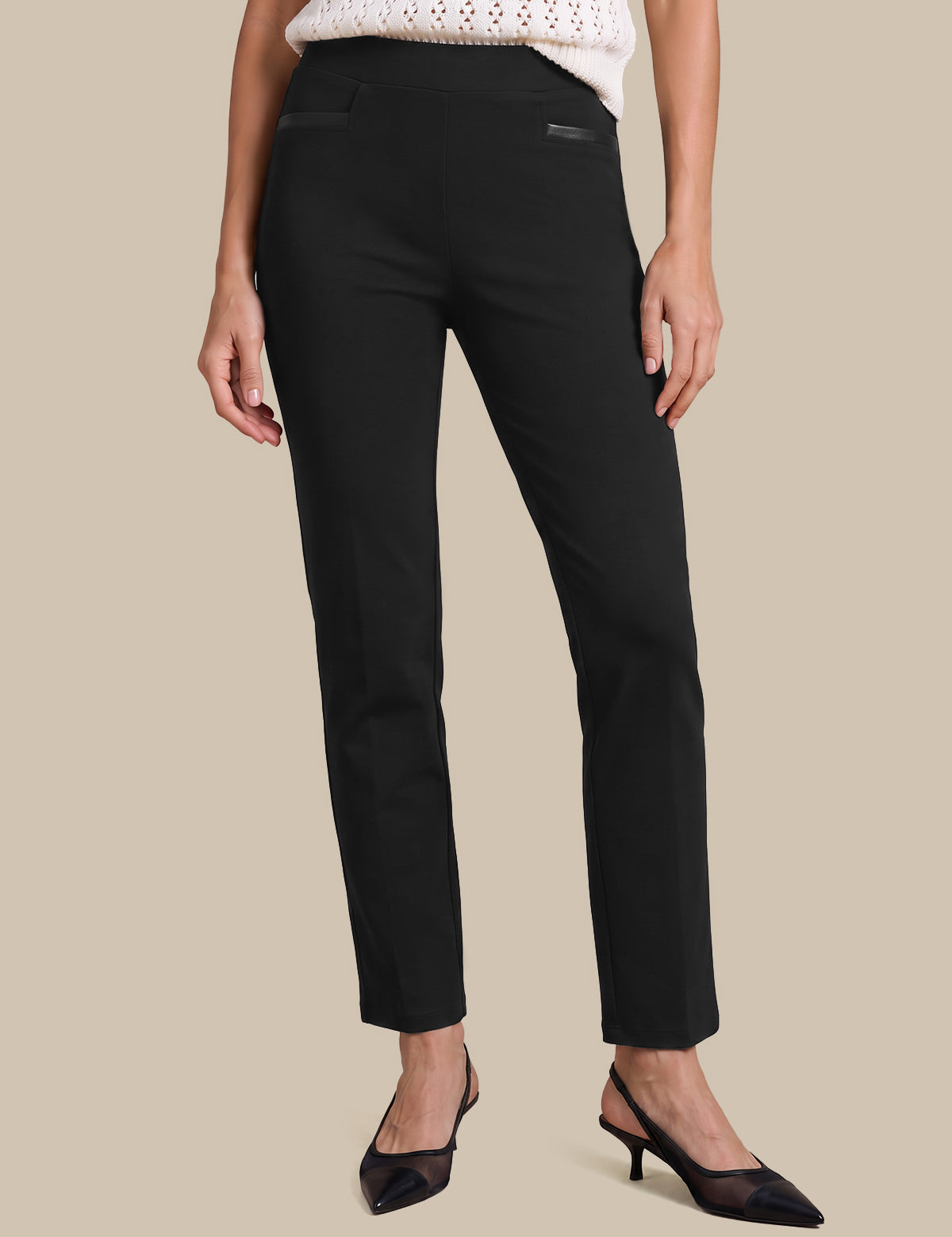 A model wearing black ankle-length straight-leg trousers, styled with a white knit top and black pointed-toe kitten heels, set against a solid beige background.