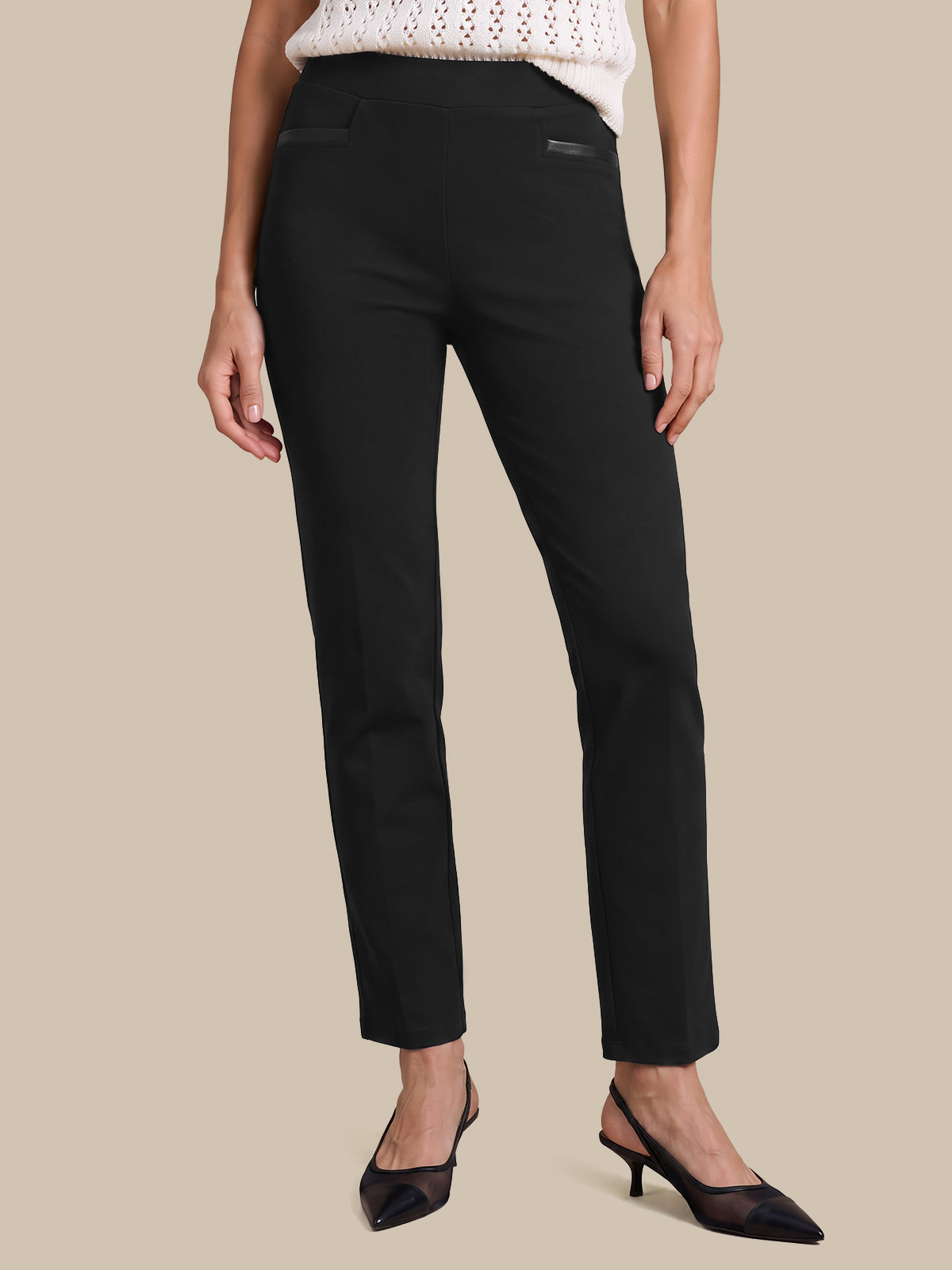 A model wearing black ankle-length straight-leg trousers, styled with a white knit top and black pointed-toe kitten heels, set against a solid beige background.