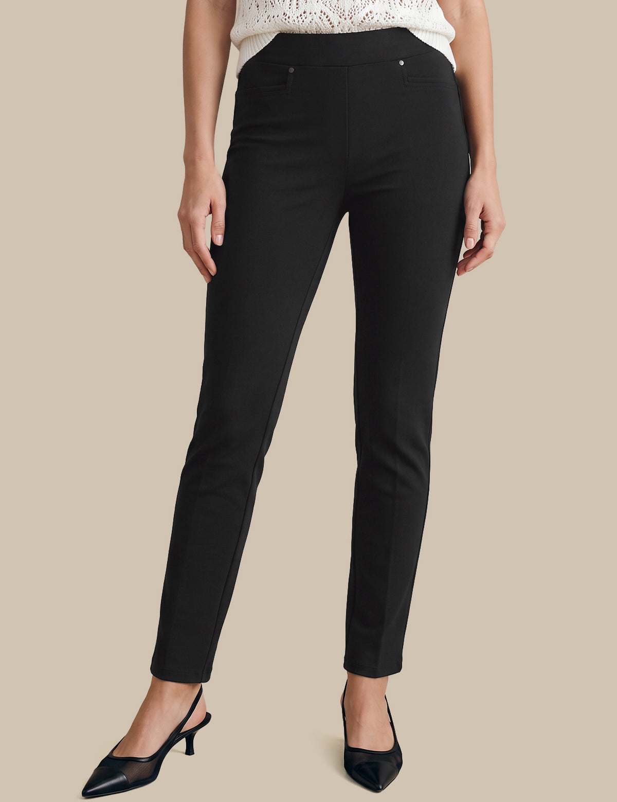 A model wearing slim-fit black ankle-length trousers, paired with a white lace-trim top and black pointed slingback heels, set against a solid beige background.