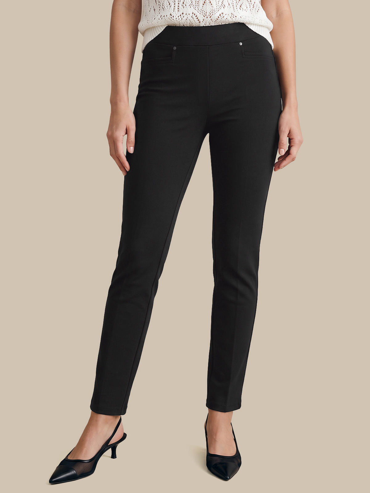 A model wearing slim-fit black ankle-length trousers, paired with a white lace-trim top and black pointed slingback heels, set against a solid beige background.