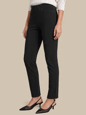 A model wearing black slim-fit ankle-length trousers, styled with a white knit top and black pointed-toe slingback heels, set against a solid beige background.