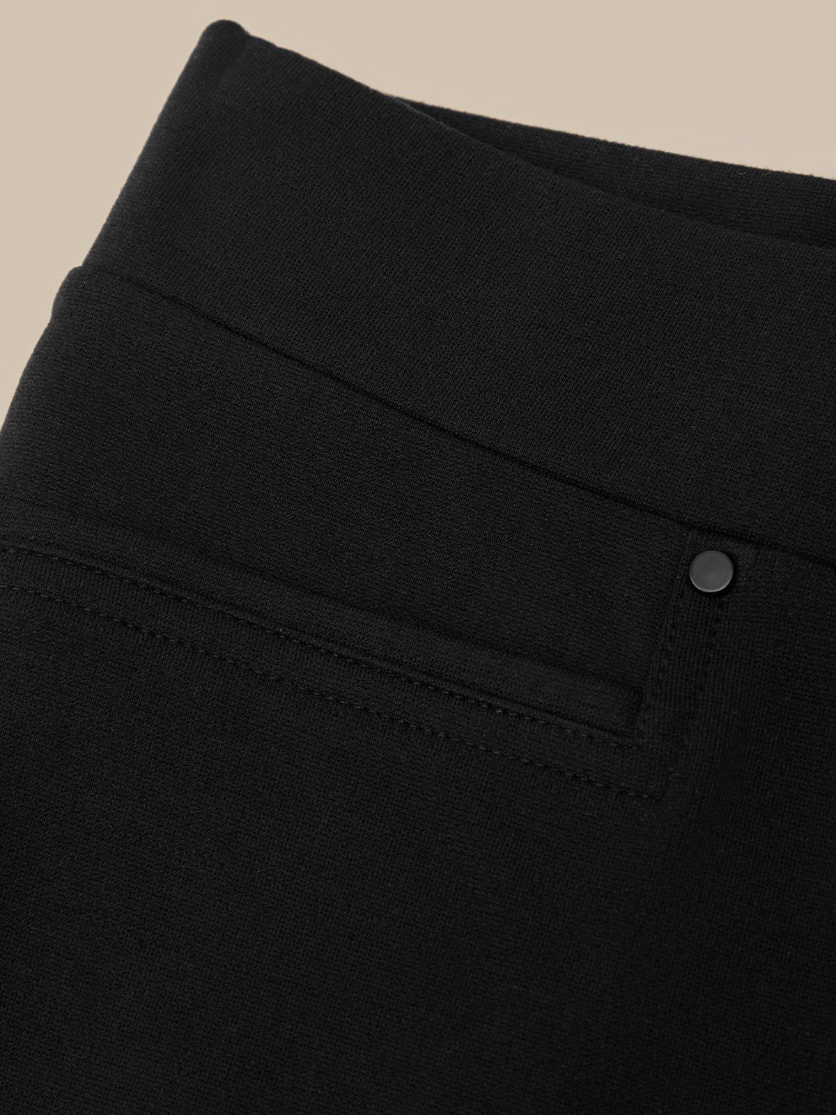Close-up of a black garment, likely a skirt or dress, showing a wide waistband, a side pocket with a button, and fine fabric texture against a light beige background.