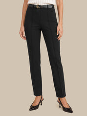 A model wearing a pair of black high-waisted, straight-leg trousers with a matching black belt, styled with a white top and black pointed-toe heels against a solid beige background.