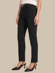 A model wearing black slim-fit ankle-length trousers with a matching black belt, paired with black slingback heels and a white knit top, set against a solid beige background.
