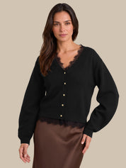 A woman modeling a black cardigan with gold buttons and a black lace V-neck trim, paired with a brown satin skirt, against a solid beige background.
