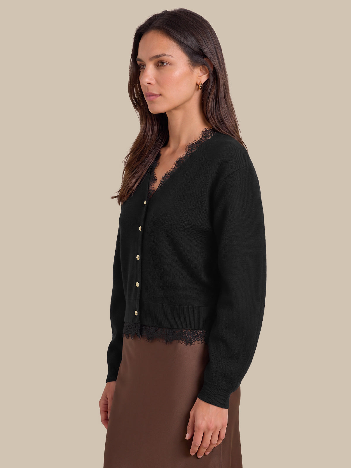 A woman wearing a black cardigan with delicate lace trim along the V-neck and hem, paired with a brown skirt, photographed against a solid beige background.