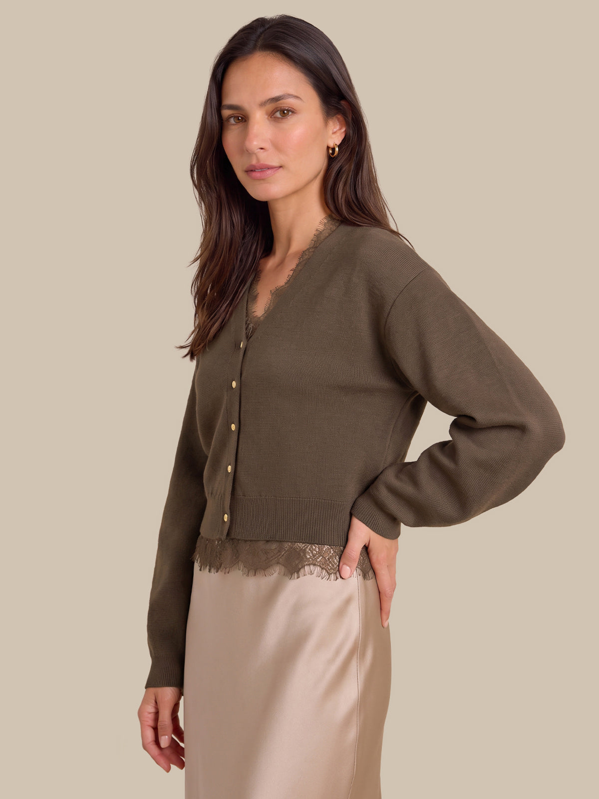 A woman modeling a cropped olive green knit cardigan with gold buttons and a lace hem, paired with a beige satin skirt, against a solid beige background.