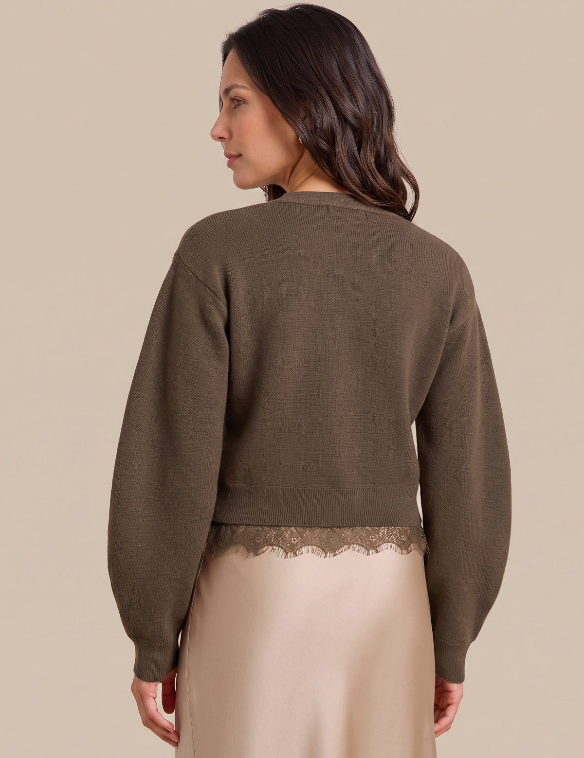 A woman shown from the back wearing a cropped, long-sleeve knit sweater in a muted taupe-brown color, paired with a light beige satin skirt featuring a lace hem. The background is a solid, warm beige studio backdrop.