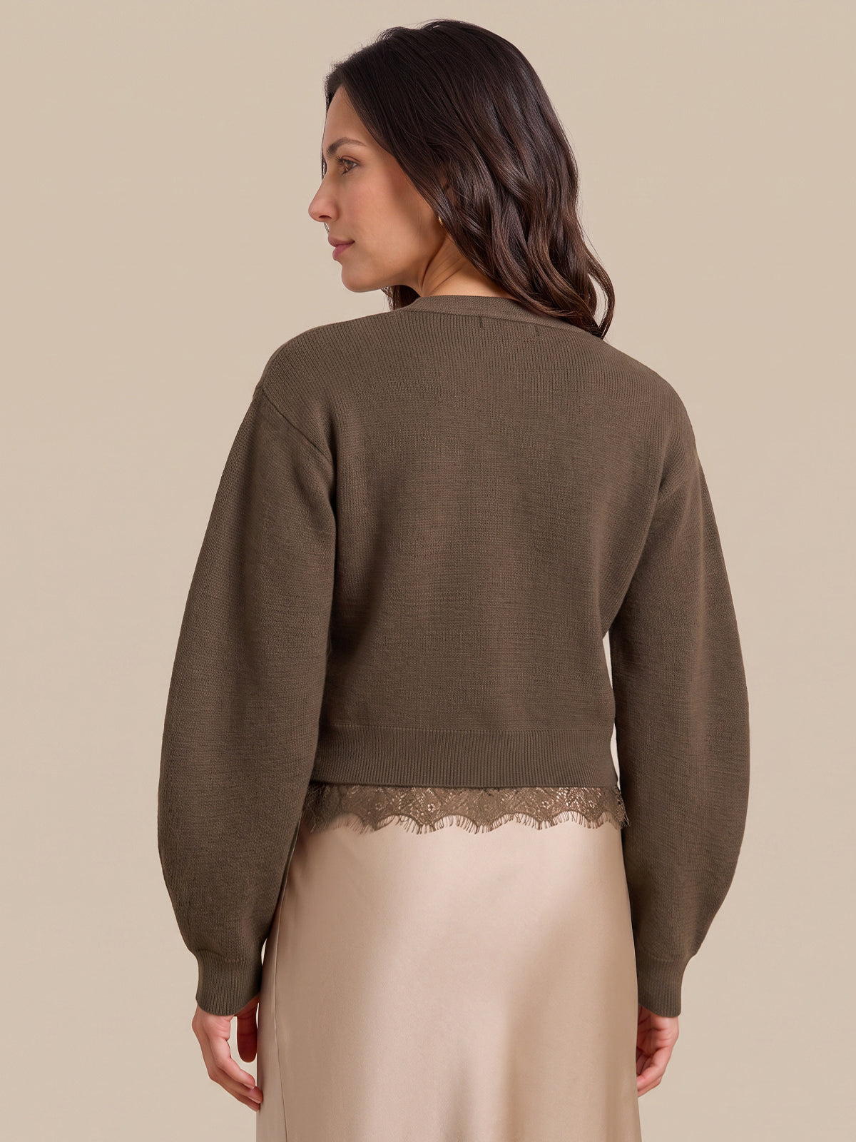 A woman shown from the back wearing a cropped, long-sleeve knit sweater in a muted taupe-brown color, paired with a light beige satin skirt featuring a lace hem. The background is a solid, warm beige studio backdrop.