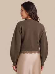 A woman shown from the back wearing a cropped, long-sleeve knit sweater in a muted taupe-brown color, paired with a light beige satin skirt featuring a lace hem. The background is a solid, warm beige studio backdrop.