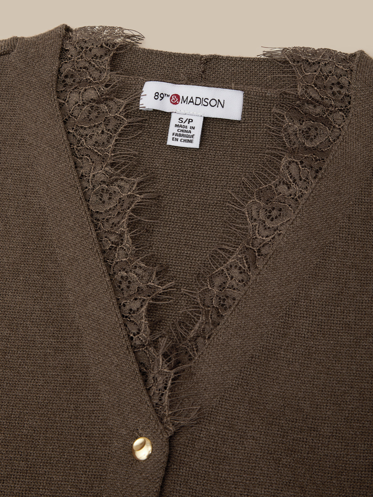 Close-up of a brown knit cardigan with a V-neck and floral lace trim, showing the interior neck label that reads 89° MADISON.