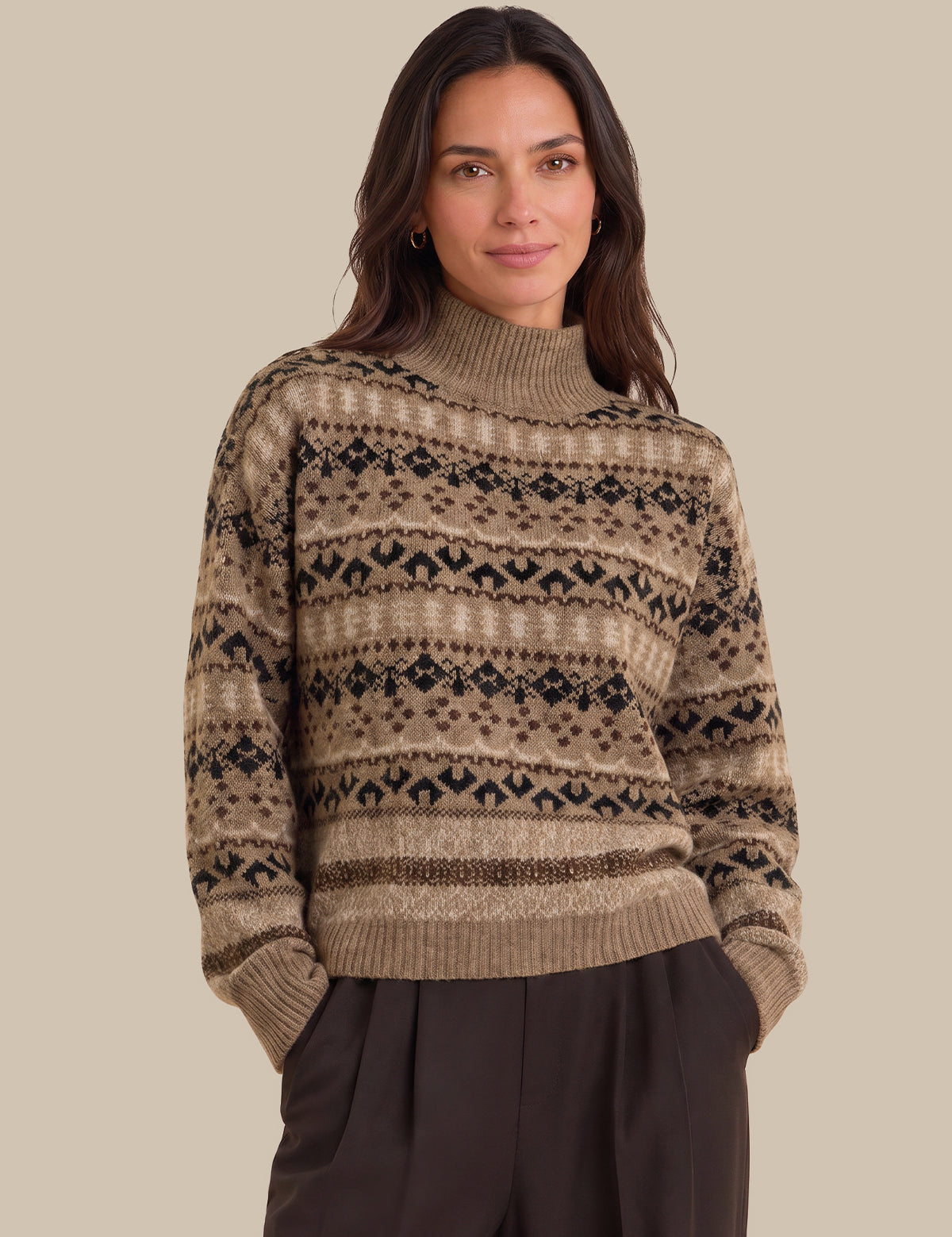 A woman wearing a beige and brown Fair Isle patterned knit sweater with a mock neck, paired with dark brown trousers, set against a solid beige background.