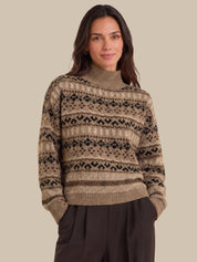 A woman wearing a beige and brown Fair Isle patterned knit sweater with a mock neck, paired with dark brown trousers, set against a solid beige background.