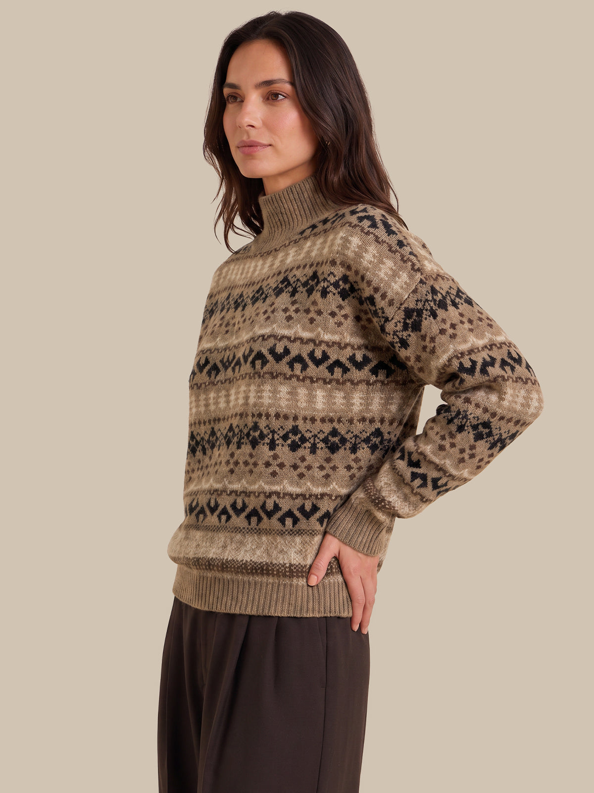 A woman wearing a beige and brown Fair Isle patterned turtleneck sweater, paired with dark brown trousers, standing against a solid beige background.
