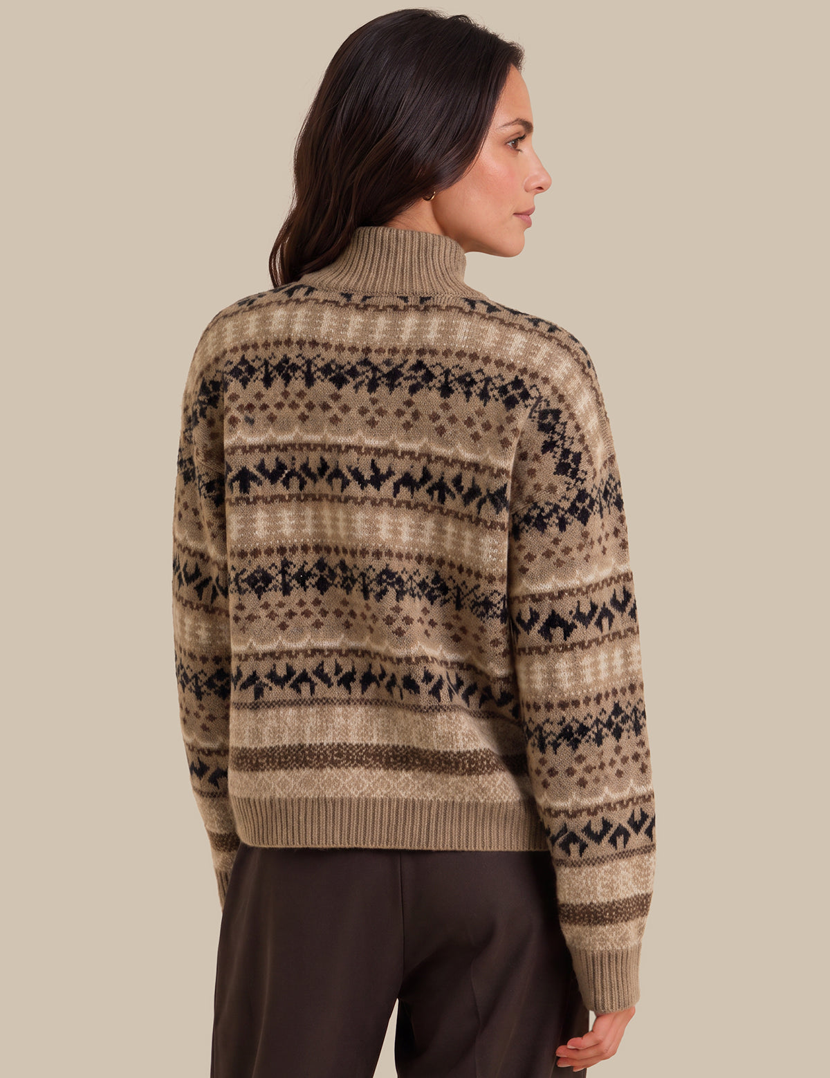 A woman shown from the back wearing a beige and brown Fair Isle patterned knit sweater with dark brown trousers, set against a solid beige studio background.