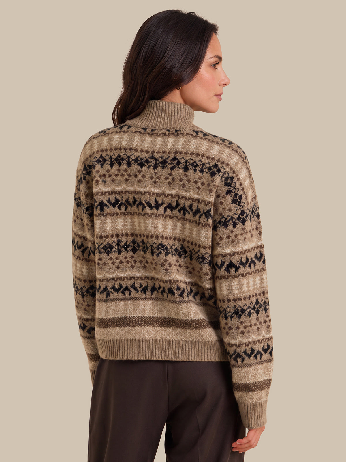 A woman shown from the back wearing a beige and brown Fair Isle patterned knit sweater with dark brown trousers, set against a solid beige studio background.