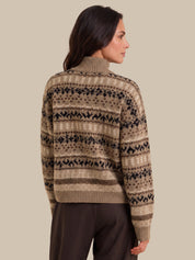 A woman shown from the back wearing a beige and brown Fair Isle patterned knit sweater with dark brown trousers, set against a solid beige studio background.