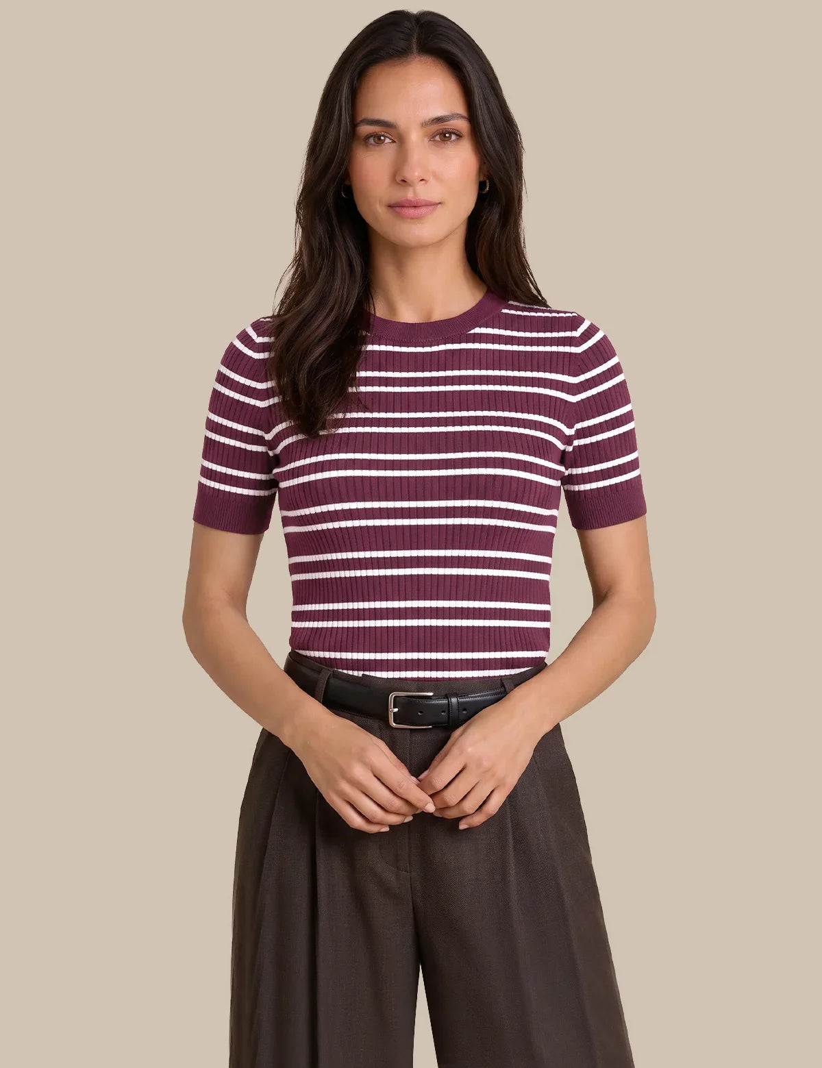 A woman modeling a short-sleeve ribbed knit top with horizontal burgundy and white stripes, paired with dark brown wide-leg trousers and a black belt, against a solid beige background.