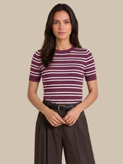 A woman modeling a short-sleeve ribbed knit top with horizontal burgundy and white stripes, paired with dark brown wide-leg trousers and a black belt, against a solid beige background.
