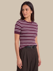 A woman modeling a short-sleeve ribbed knit top with horizontal burgundy and white stripes, paired with dark brown pleated trousers, against a solid beige background.