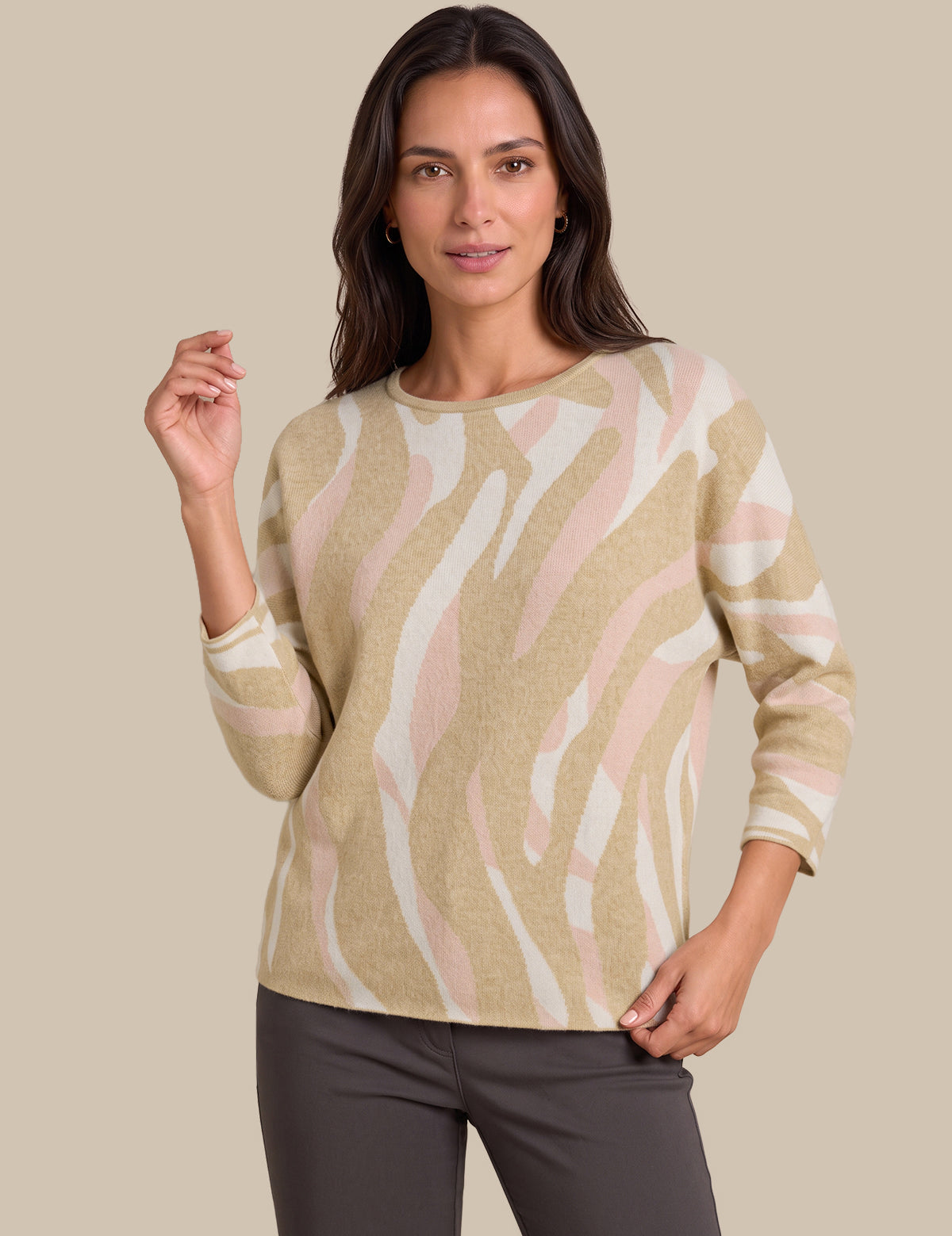 A woman modeling a beige, white, and light pink zebra-patterned knit sweater with three-quarter sleeves, paired with dark gray pants, against a solid beige background.